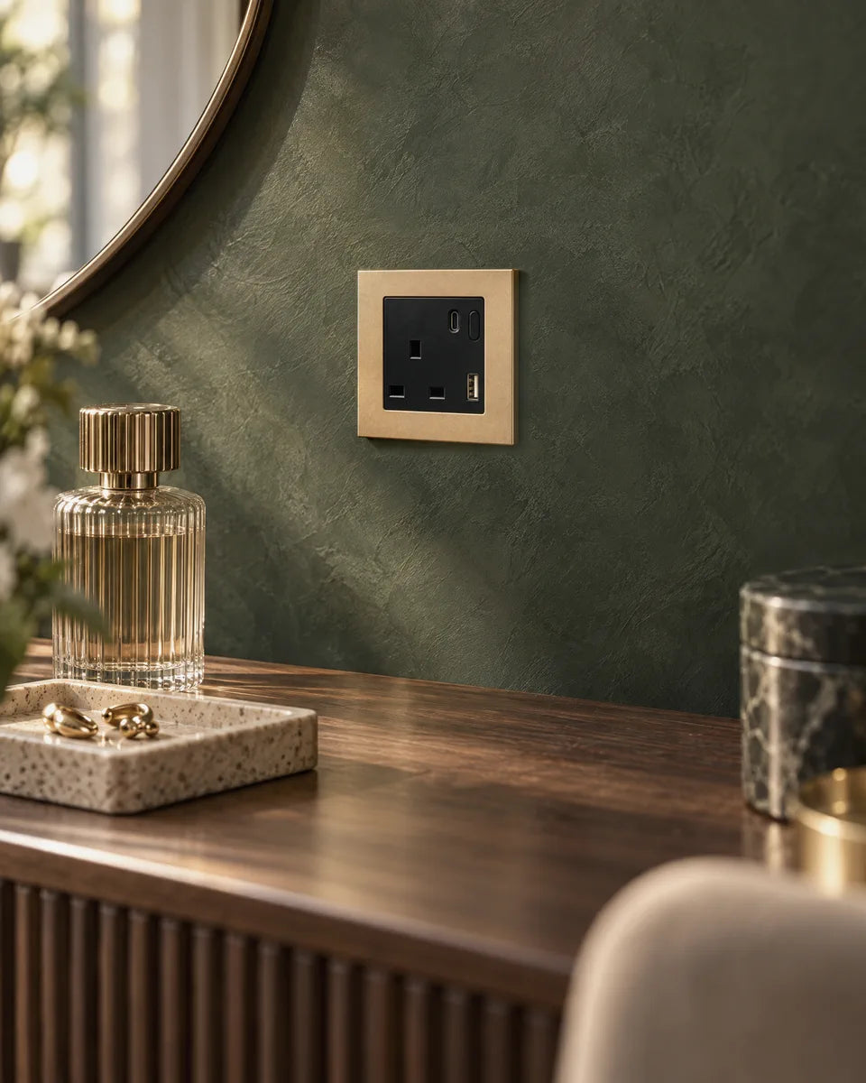 A gold aluminium smart socket on a deep green plaster wall above a walnut dressing table