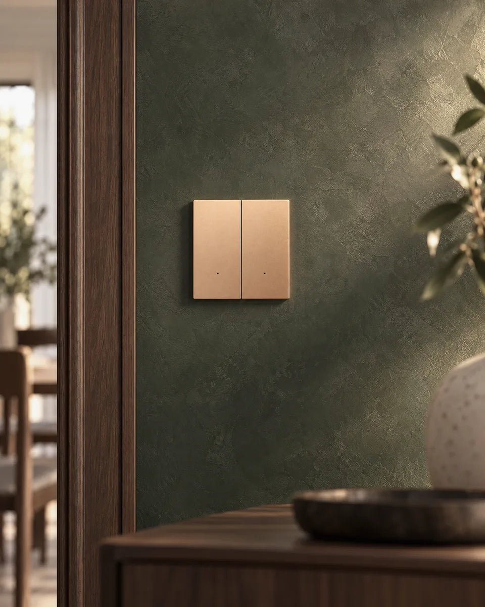 A gold aluminium 2-gang smart light switch on a deep green plaster wall beside a walnut door frame