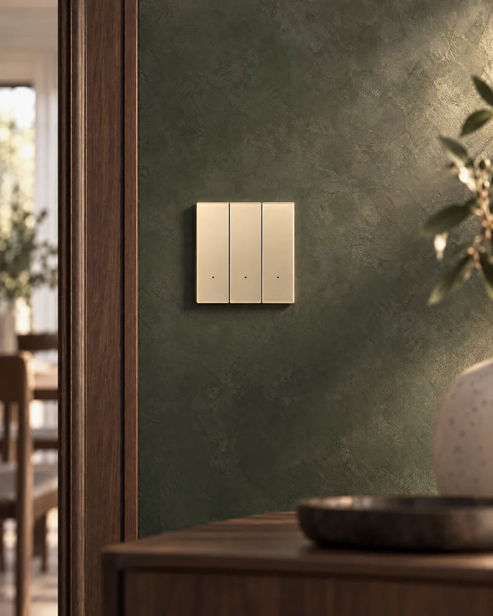 A gold glass 3-gang smart light switch on a deep green plaster wall beside a walnut door frame