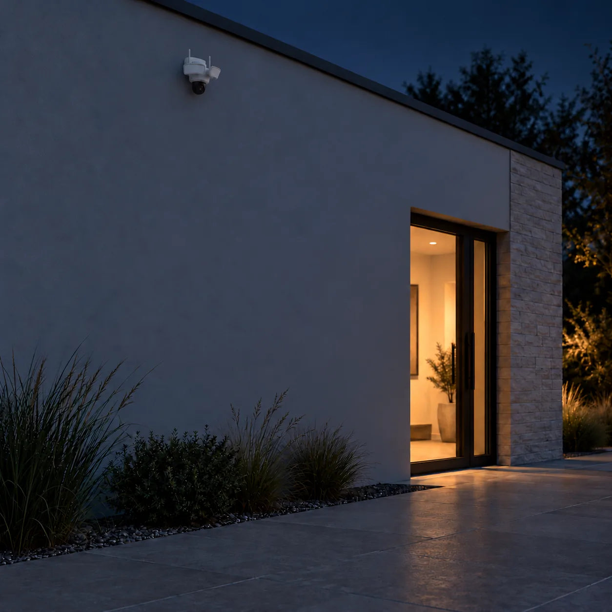 Floodlight camera Pro 5MP fitted high under the roofline of a house at night, its lights switching on over the planting and paving